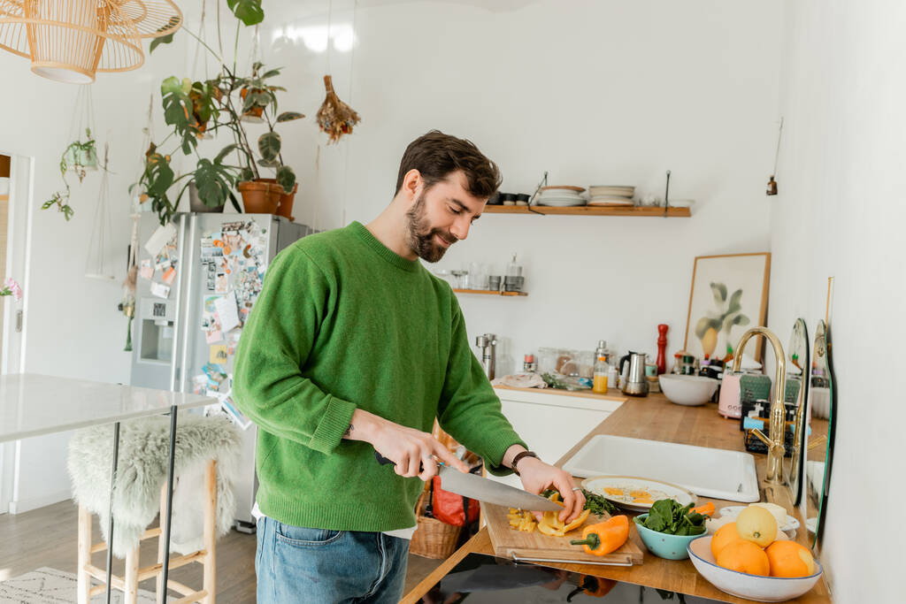 キッチンで朝食を調理しながら新鮮な唐辛子を切るジャンパーとジーンズの幸せなひげ付き男  - 写真・画像