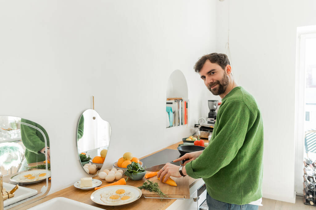 現代のキッチンで朝食を調理しながら新鮮な唐辛子を切る緑のジャンパーの幸せな男  - 写真・画像