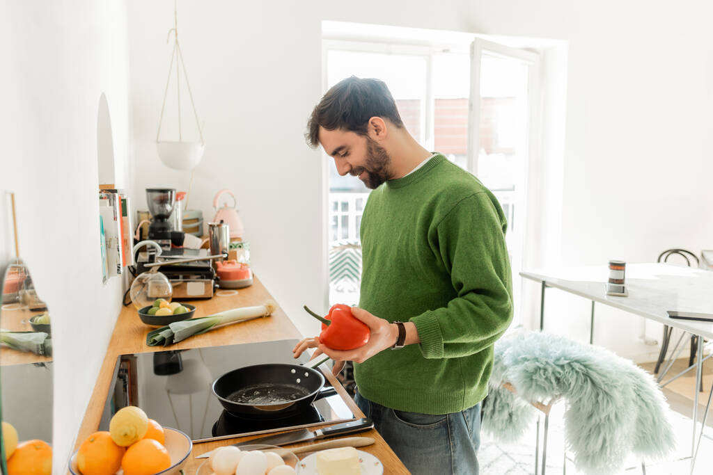 позитивний бородатий чоловік тримає болгарський перець біля сковороди під час приготування сніданку вдома  - Фото, зображення