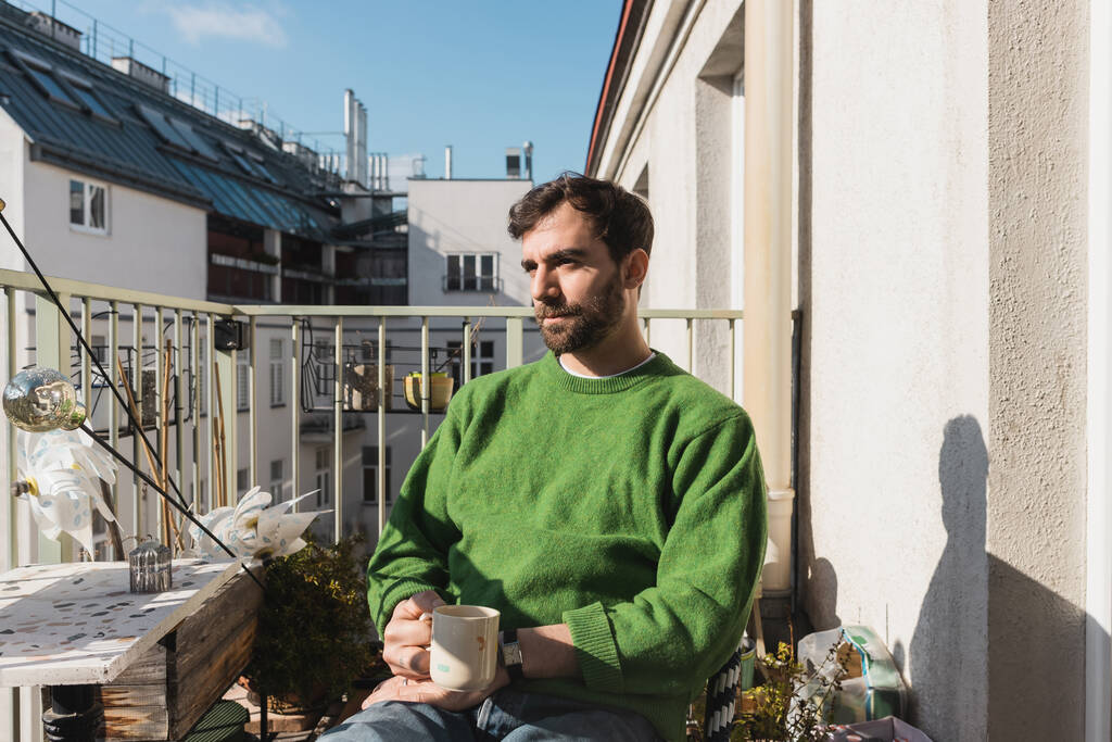 hombre relajado en jersey verde y jeans sosteniendo taza de café en la mañana en el balcón - Foto, Imagen