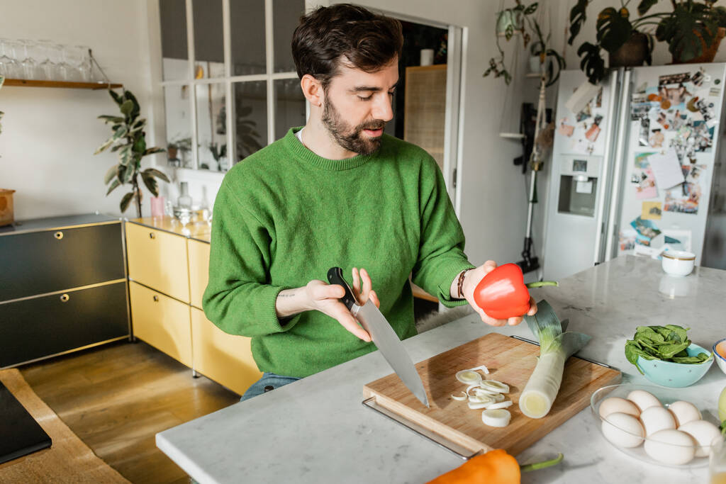 現代のキッチンで調理している間,ナイフとベルペッパーを保持する緑のジャンパーのひげ付きタトゥー男 - 写真・画像