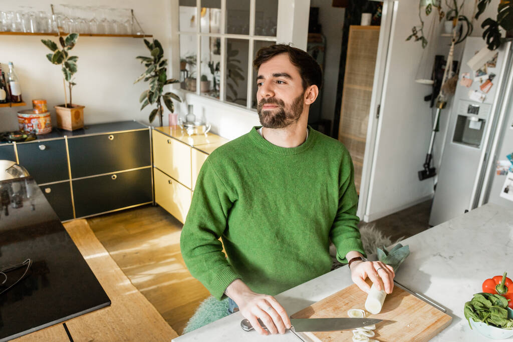Relaxed and bearded man in green jumper looking away while holding knife and leek at home  - Photo, Image