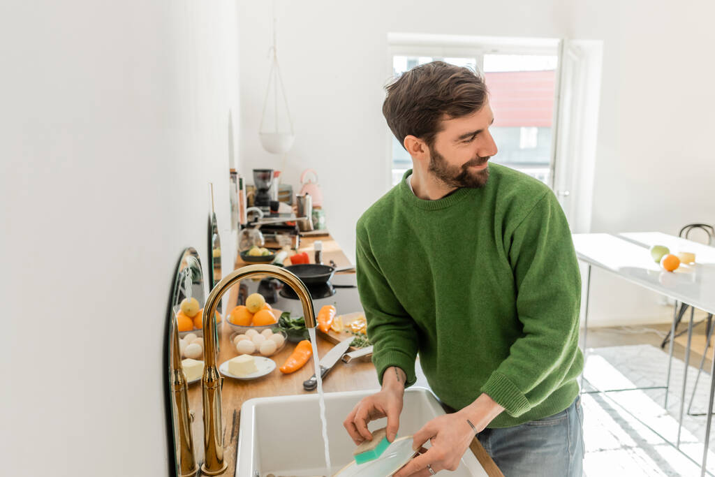 Uśmiechnięty brodaty i wytatuowany mężczyzna myjący talerz w pobliżu zlewu i patrzący w domu  - Zdjęcie, obraz