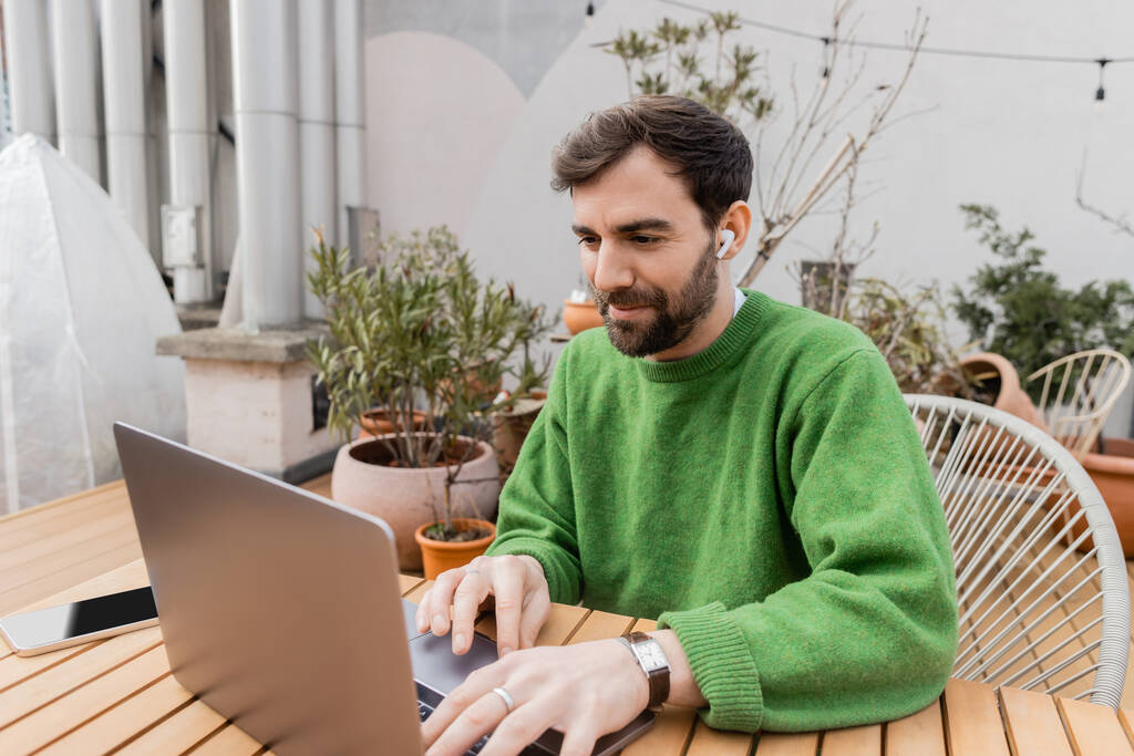 фрилансер в зеленом свитере с помощью наушника и ноутбука во время работы на крыше дома терраса  - Фото, изображение