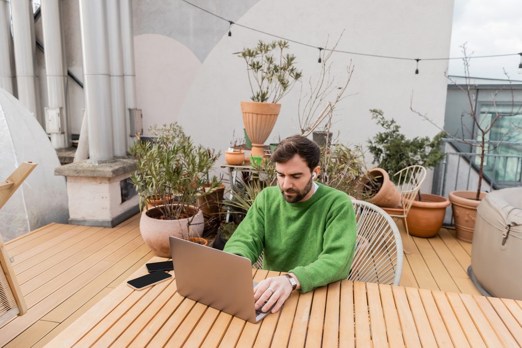 empreendedor em fone de ouvido sem fio usando laptop enquanto trabalhava perto de smartphones no terraço da casa  - Foto, Imagem