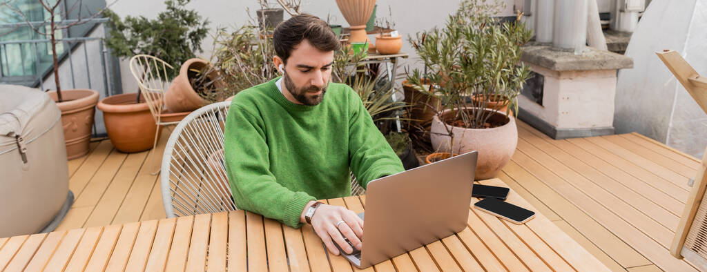 freelancer in earphone using laptop and working on rooftop terrace of house, banner  - Photo, Image