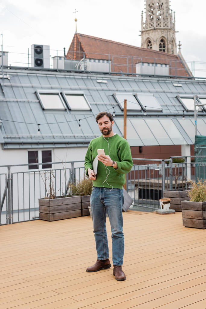 freelancer using earphones and smartphone and standing on rooftop terrace in Vienna  - Photo, Image