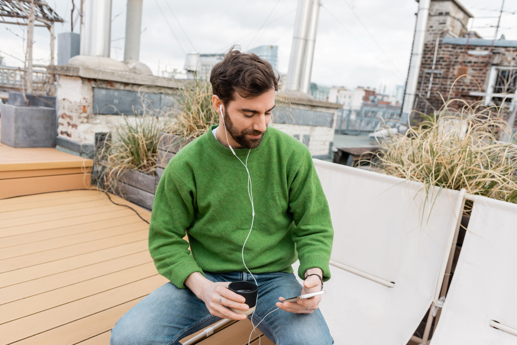 vousatý muž poslouchající hudbu ve sluchátkách a držící šálek kávy na střeše ve Vídni, Rakousko - Fotografie, Obrázek