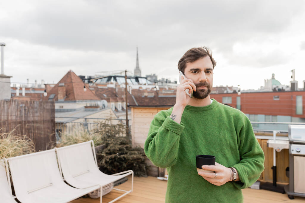 グリーンジャンパーの男性は,スマートフォンで話し,オーストリアのウィーンで屋上にコーヒーを飲みます - 写真・画像