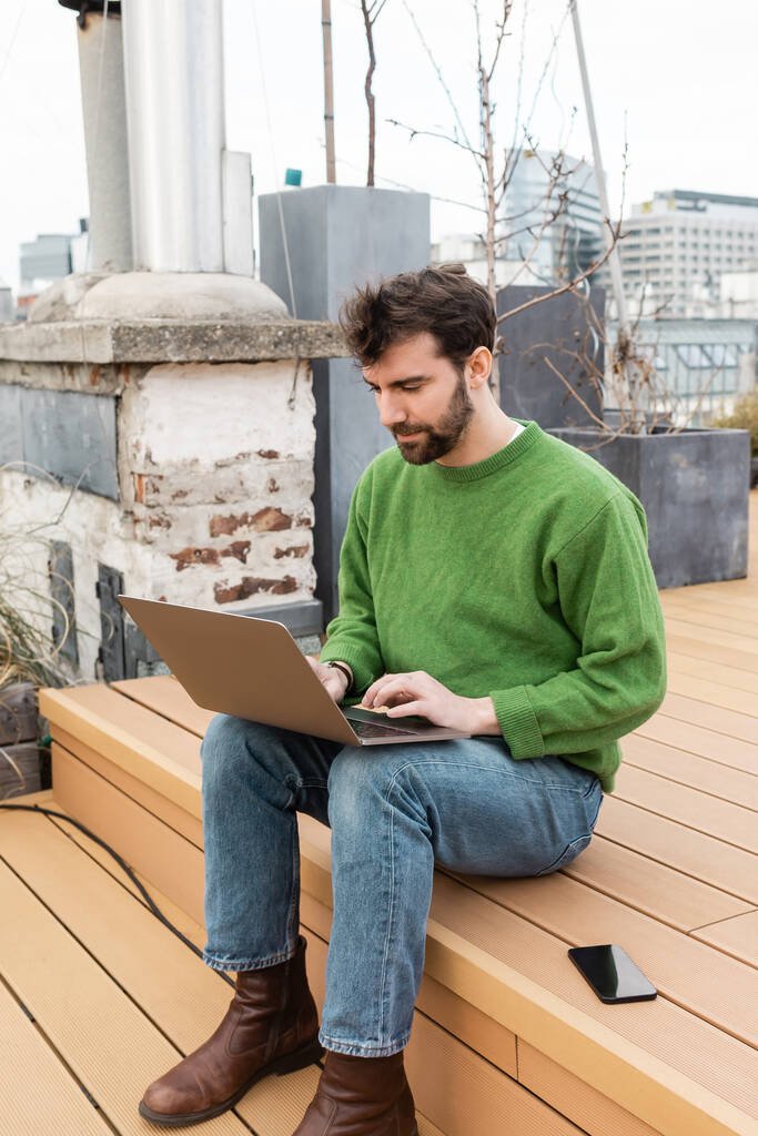 freelancer bonito usando laptop perto de smartphone enquanto trabalhava no telhado em Viena, Áustria - Foto, Imagem