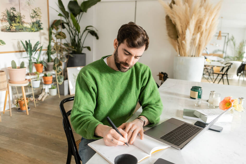 Γενειοφόρος ελεύθερος επαγγελματίας σε πράσινο jumper γράφοντας στο σημειωματάριο κοντά συσκευές, φλιτζάνι καφέ και κεριά  - Φωτογραφία, εικόνα