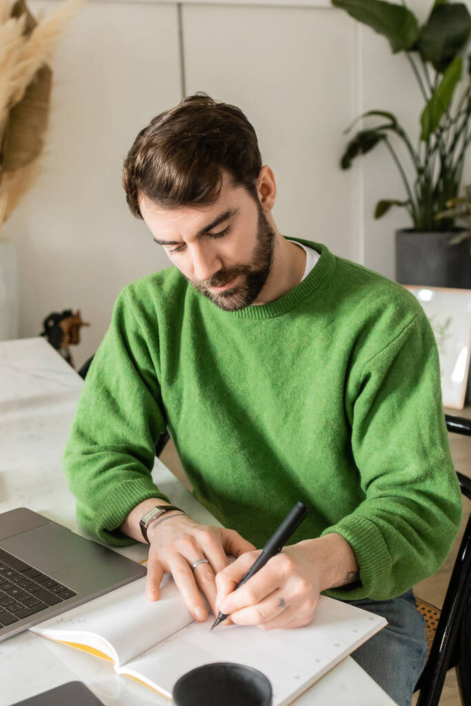 freelancer barbudo en jersey verde escrito en portátil cerca de laptop, smartphone y taza de café - Foto, imagen