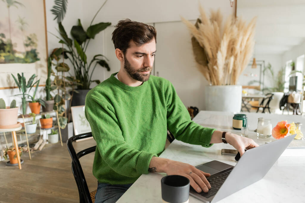 Γενειοφόρος και μελαχρινή ελεύθερος επαγγελματίας σε πράσινο άλτης χρησιμοποιώντας φορητό υπολογιστή και τη λήψη smartphone στο τραπέζι - Φωτογραφία, εικόνα