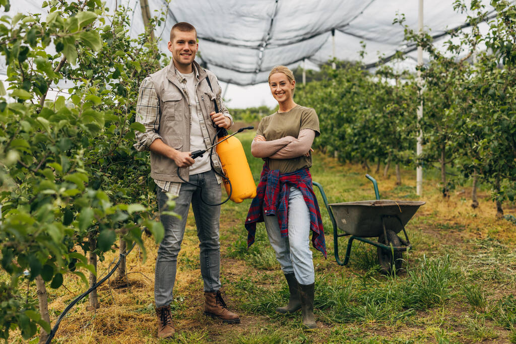 Ein Mann hält einen Sprayer in der Hand, neben ihm steht eine Frau in einem Obstbauernhof. - Foto, Bild