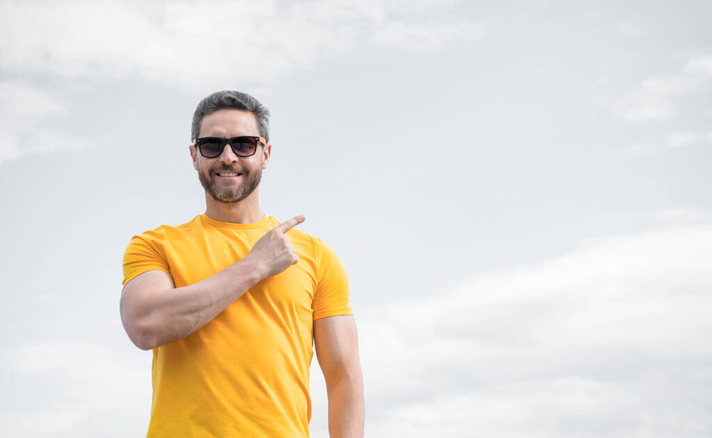 ευτυχισμένος άνθρωπος σε κίτρινο πουκάμισο και γυαλιά ηλίου δείχνουν δάχτυλο στο φόντο του ουρανού. - Φωτογραφία, εικόνα