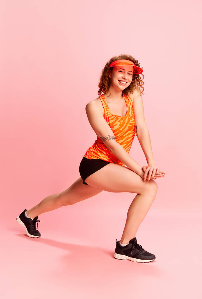 Retrato de cuerpo entero de una joven en entrenamiento de ropa deportiva, haciendo ejercicios, estirándose sobre fondo de estudio rosa. Concepto de emociones humanas, cultura juvenil, moda, estilo de vida deportivo - Foto, Imagen