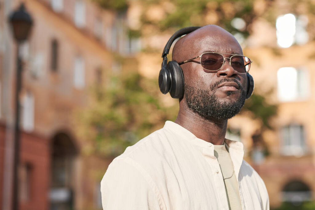 Портрет серьезного крутого черного парня с бородой и лысой головой, стоящего в городе и слушающего музыку в наушниках - Фото, изображение