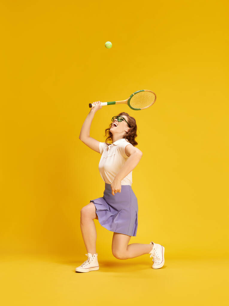 Portret młodej, aktywnej, ładnej dziewczyny w odzieży sportowej, tenisa grającego w ruchu uderzającego piłką z rakietą na żółtym tle studia. Pojęcie sportu, aktywnego stylu życia, emocji, mody, hobby - Zdjęcie, obraz