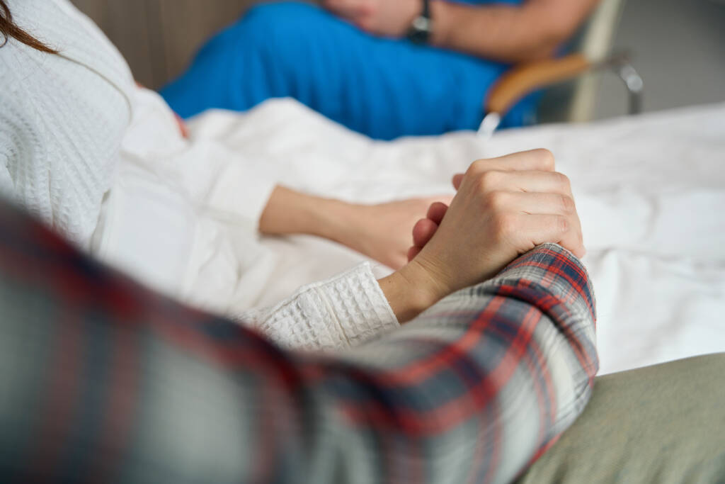 Gedeeltelijk zicht op twee personen op ziek bed die elkaars hand binnenshuis vasthouden - Foto, afbeelding