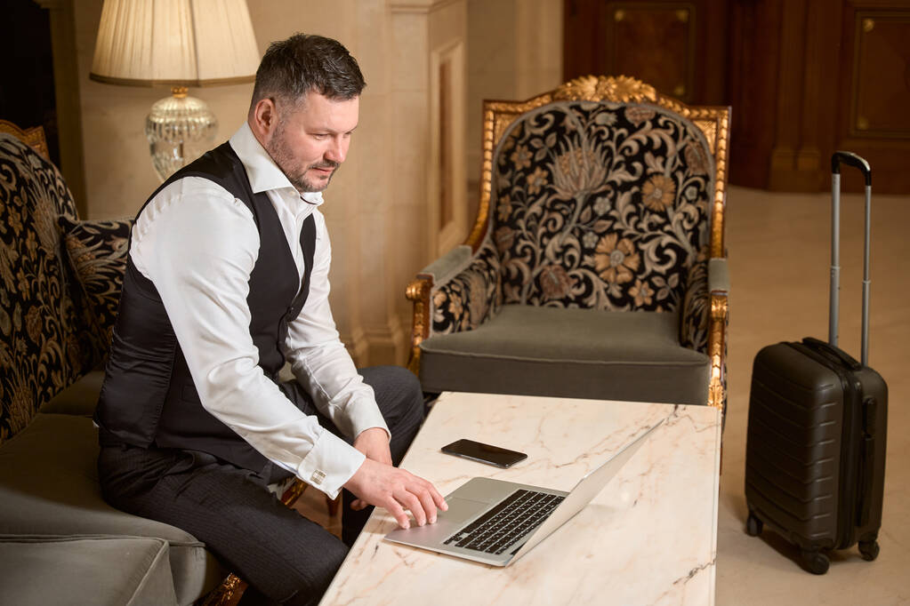 Gut aussehender seriöser Geschäftsmann arbeitet am Laptop am Tisch neben dem Gepäck in der Lobby des Hotels - Foto, Bild