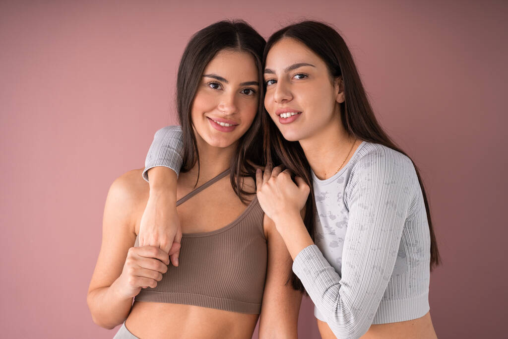 Mooie jonge vrouwen die samen poseren, glimlachen en naar de camera kijken bij roze pastel studio achtergrond. Schoonheidsportret van twee vrouwelijke modellen. Perfecte glimlach - Foto, afbeelding
