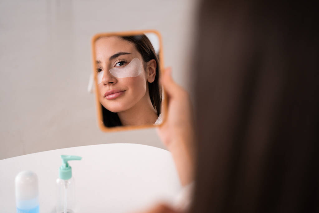 Heijastus peilistä söpö brunette nainen laastaria silmien alla. Rauhallinen nainen seisoo kylpyhuoneessa ja etsii peili - Valokuva, kuva