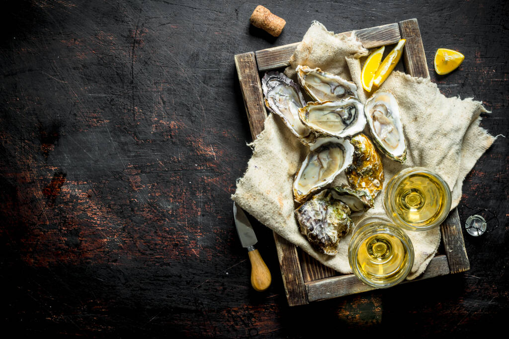 白ワインのグラスと木製のトレイのぼろに生の牡蠣。暗い素朴な背景に - 写真・画像