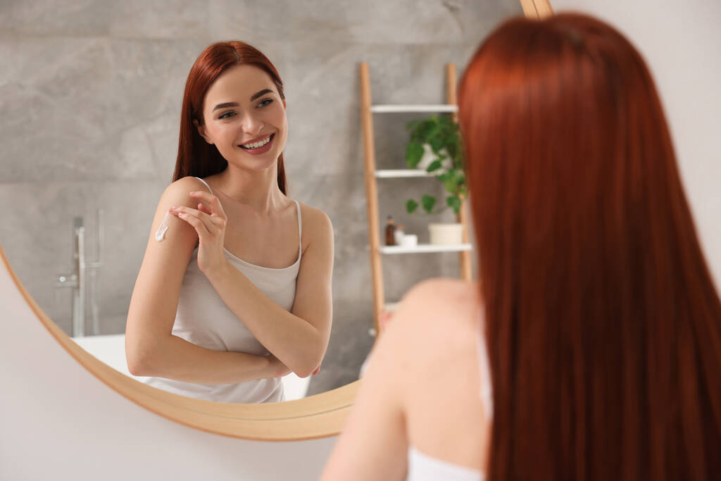 Gyönyörű fiatal nő alkalmazó testápoló krém vállára közel tükör a fürdőszobában - Fotó, kép