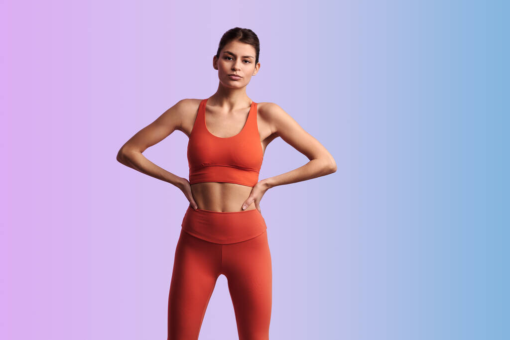 Young confident Hispanic female athlete in red tight activewear standing with hands on waist against gradient background during fitness workout - Photo, Image
