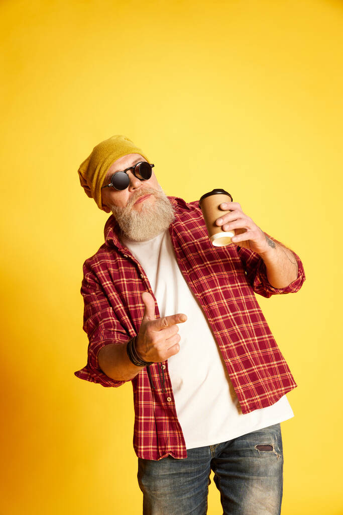 Retrato de hombre barbudo maduro en ropa elegante, camisa a cuadros, gafas y sombrero, posando con café sobre fondo amarillo estudio. Concepto de emociones humanas, estilo de vida, moda masculina, estilo - Foto, imagen