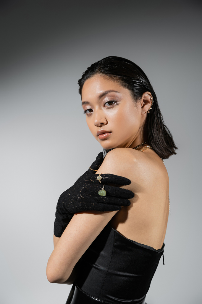 brünette und asiatische junge Frau mit kurzen Haaren posiert in schwarzem trägerlosen Kleid und Handschuhen mit Goldschmuck vor der Kamera auf grauem Hintergrund, nasse Frisur, Halsketten, Ohrringe, natürliches Make-up - Foto, Bild