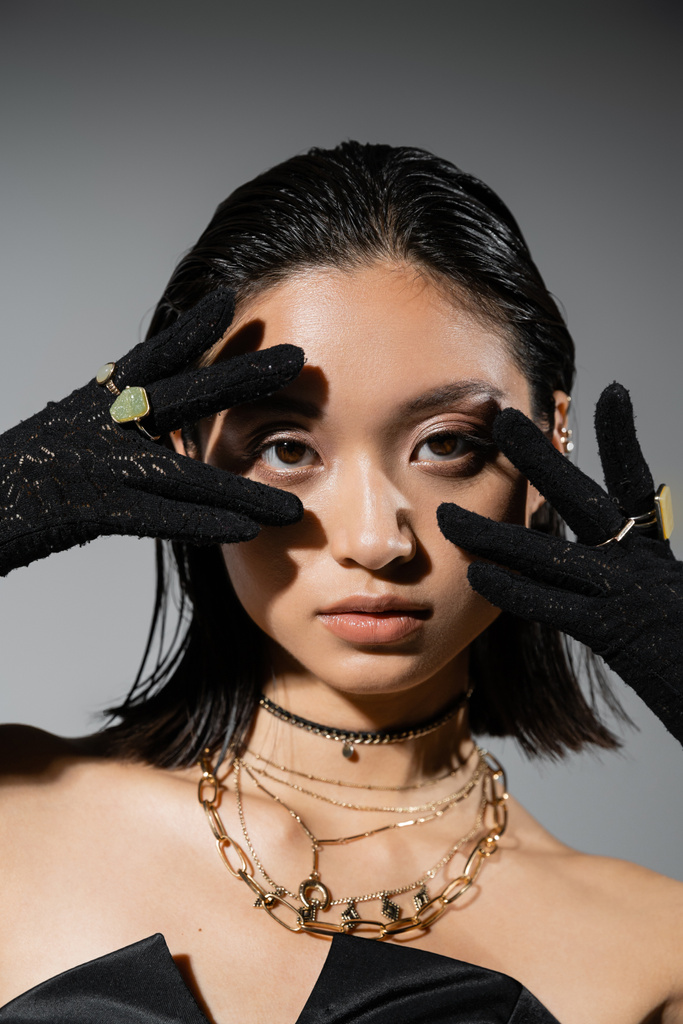 retrato de morena e asiática jovem mulher com cabelo curto posando em luvas pretas com anéis dourados, olhando para a câmera em fundo cinza, penteado molhado, mãos perto do rosto, maquiagem natural - Foto, Imagem