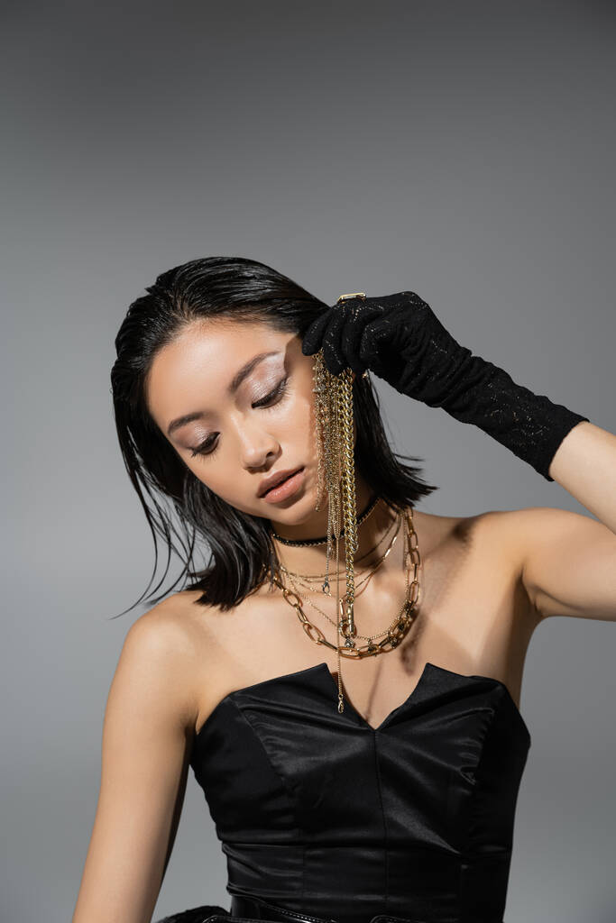 Porträt einer brünetten und asiatischen jungen Frau mit kurzen Haaren, die in schwarzen Handschuhen und trägerlosem Kleid posiert, während sie Goldschmuck auf grauem Hintergrund hält, nasse Frisur, natürliches Make-up - Foto, Bild