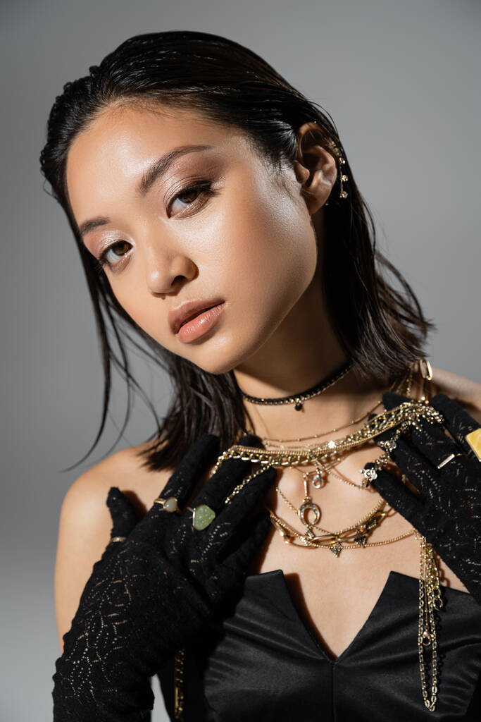 portrait de jeune femme brune et asiatique aux cheveux courts posant en gants noirs avec anneaux et robe bustier tout en tenant des bijoux dorés sur fond gris, coiffure mouillée, maquillage naturel - Photo, image