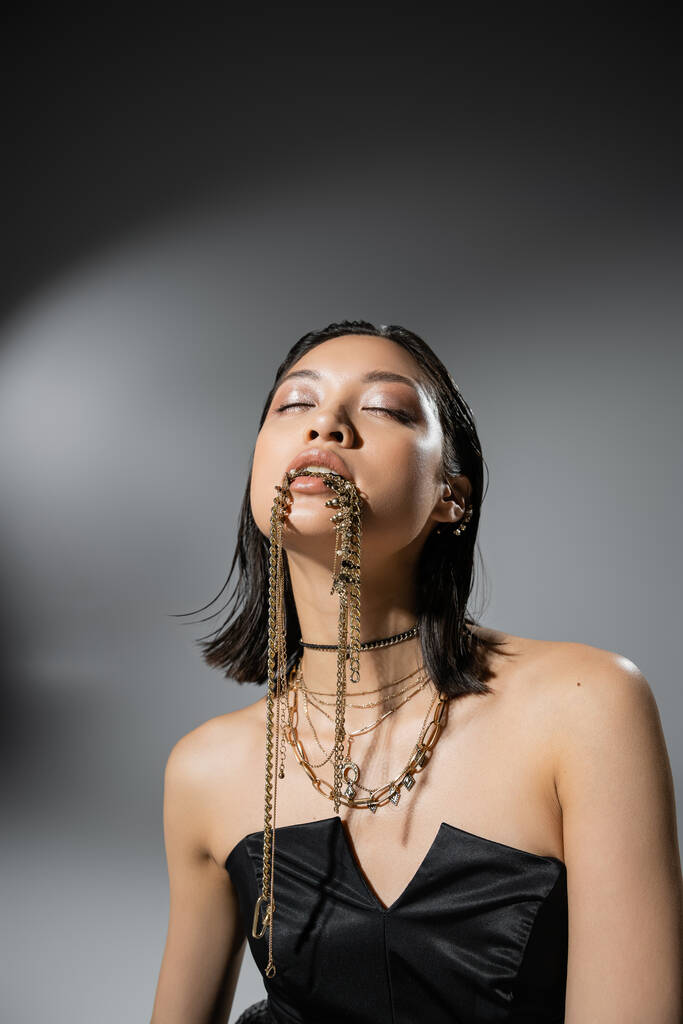 портрет азиатской молодой женщины с короткими волосами и закрытыми глазами, позирующей в черном платье без бретелек, держа золотые украшения во рту на сером фоне, влажная прическа, натуральный макияж - Фото, изображение
