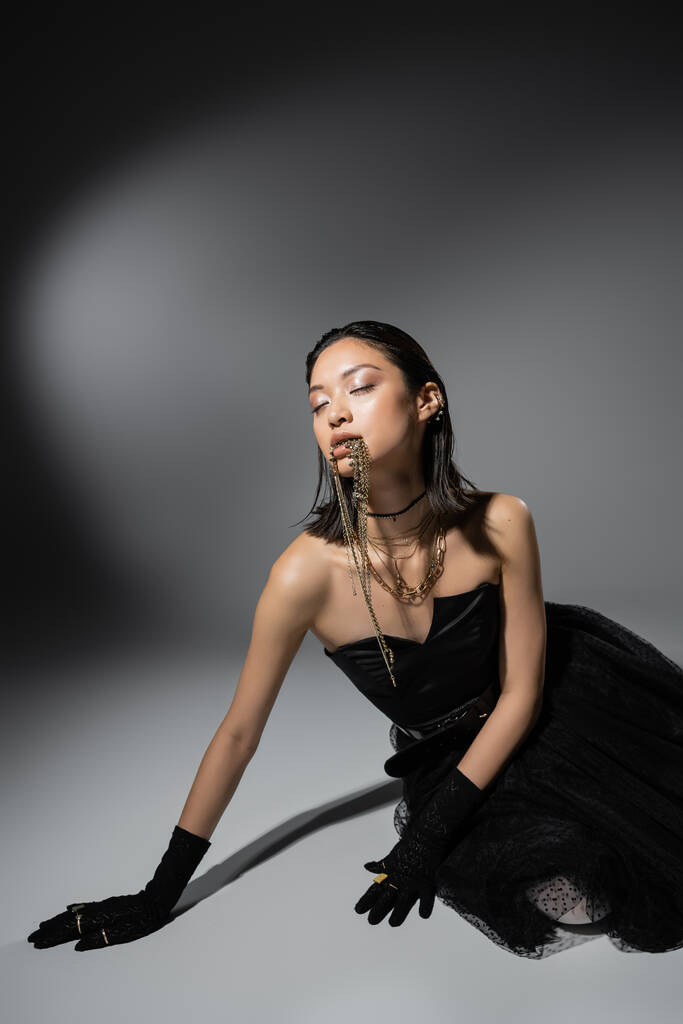 mujer joven asiática de moda con pelo corto posando en vestido negro sin tirantes y guantes mientras sostiene joyas de oro en la boca sobre fondo gris, maquillaje natural, peinado húmedo, ojos cerrados  - Foto, imagen