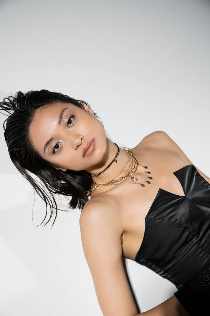 alluring and asian young woman with short hair lying in black strapless dress while posing in golden jewelry on grey background, wet hairstyle, natural makeup, looking at camera  - Photo, Image