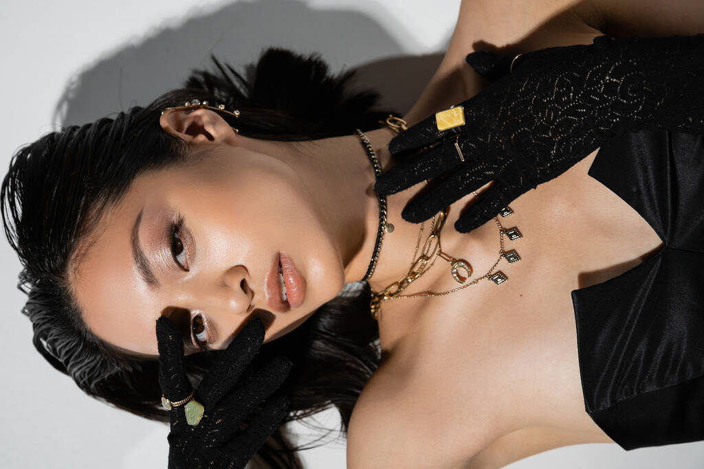 top view of young asian woman with closed eyes and short brunette hair lying in black gloves and strapless dress while posing in golden jewelry on grey background, wet hairstyle, hand near face  - Photo, Image