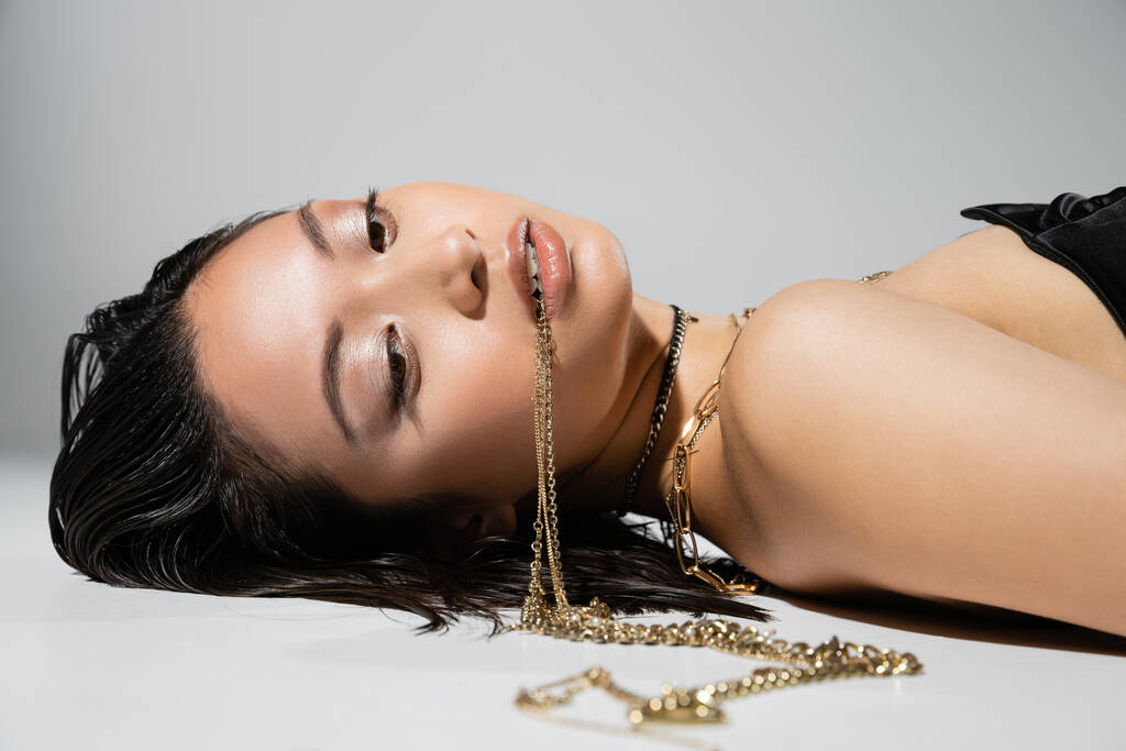 jonge aziatische vrouw met kort brunette haar met gouden sieraden in de mond, terwijl poseren en liggen op grijze achtergrond, dagelijkse make-up, nat haar, bruine ogen - Foto, afbeelding