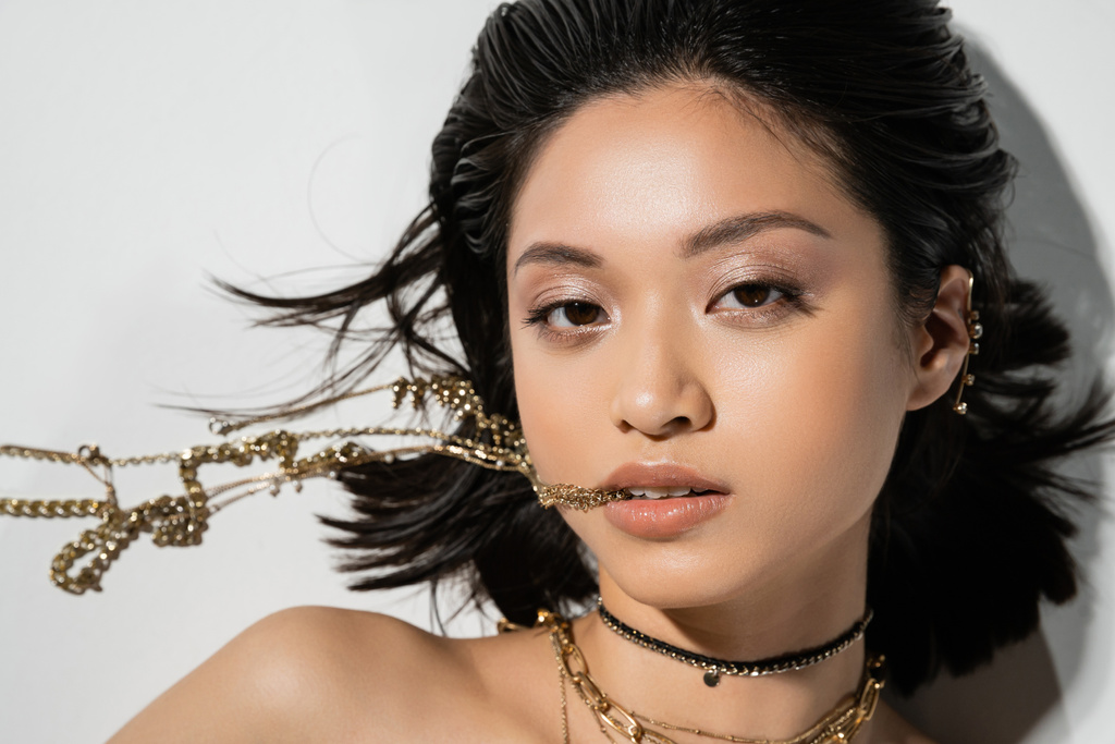 shora pohled na mladou asijskou ženu s krátkými brunetkami vlasy drží zlaté šperky v ústech při pohledu na kameru a leží na šedém pozadí, každodenní make-up, mokrý účes  - Fotografie, Obrázek
