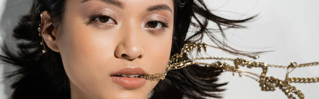jeune femme asiatique avec de courts cheveux bruns tenant des bijoux dorés dans la bouche tout en regardant la caméra et couché sur fond gris, maquillage quotidien, coiffure humide, vue supérieure, bannière  - Photo, image