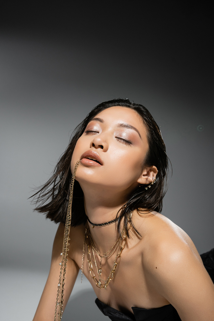 modèle asiatique élégant avec cheveux bruns courts tenant des bijoux dorés dans la bouche, posant en robe bustier sur fond gris, maquillage quotidien, coiffure humide, jeune femme, yeux fermés, portrait  - Photo, image