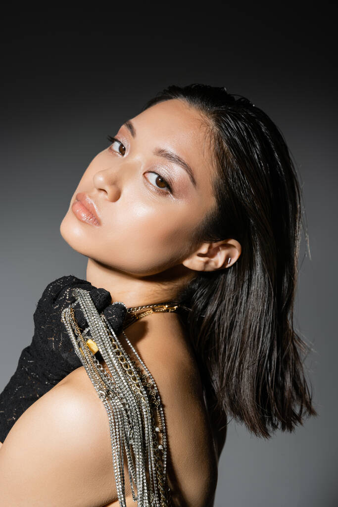 muotokuva houkutteleva aasialainen nuori nainen lyhyet hiukset poseeraa musta käsine ja tilalla kultainen ja hopea koruja olkapäällä seisoessaan harmaa tausta, märkä kampaus, luonnollinen meikki - Valokuva, kuva