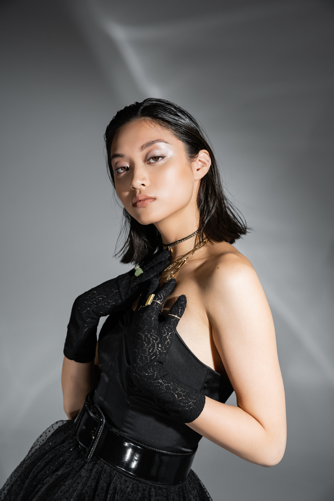 retrato de sedutor asiático jovem mulher com cabelo curto posando em preto vestido sem alças com cinto e luvas enquanto olha para a câmera no fundo cinza, penteado molhado, colares dourados  - Foto, Imagem