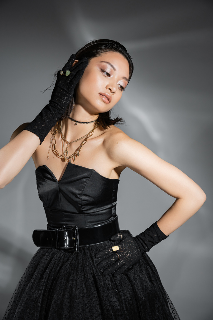 гламурная молодая азиатская женщина с короткими волосами, позирующая с рукой на бедре в черном платье без бретелек с поясом и перчатками, отворачиваясь на сером фоне, влажная прическа, золотые ожерелья  - Фото, изображение