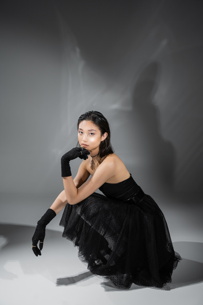 full length of stylish asian young woman with short hair sitting in black strapless dress with tulle skirt and gloves while looking at camera on grey background, wet hairstyle, golden necklaces  - Photo, Image