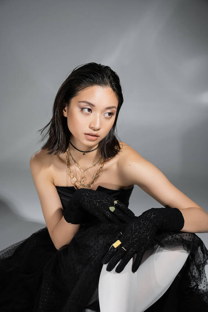 привлекательная азиатская молодая женщина с короткими волосами, сидящая в черном платье без бретелек с юбкой тюля и перчатками, смотрящая в сторону на сером фоне, влажная прическа, золотые ожерелья, мечтательная модель - Фото, изображение