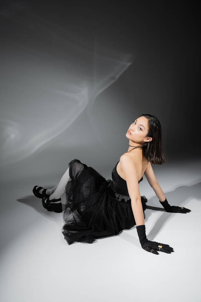 full length of asian young woman with short hair sitting in black strapless dress with tulle skirt white tights, shoes and gloves while looking at camera on grey background, wet hairstyle, captivating - Photo, Image