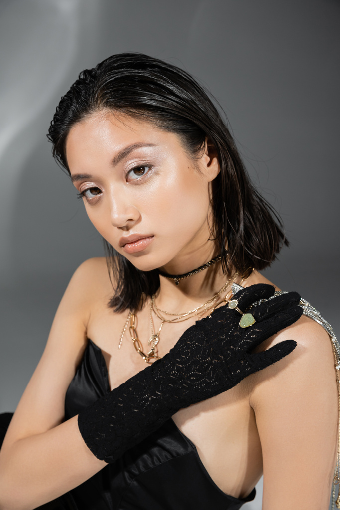 enchanting asian model with short and brunette hair holding golden jewelry in hand while posing in strapless dress and black glove on grey background, everyday makeup, wet hairstyle, young woman - Photo, Image