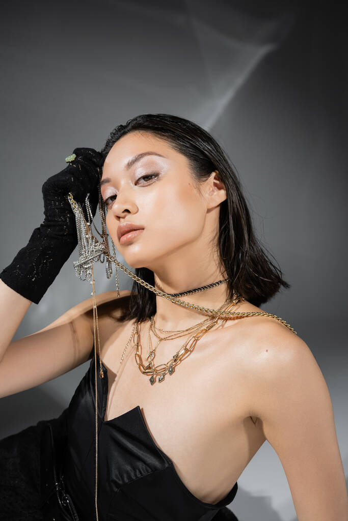 ohromující asijský model s krátkými a brunetkami vlasy drží zlaté šperky v ruce při pózování v bezramkové šaty a černé rukavice na šedém pozadí, každodenní make-up, mokré vlasy, mladá žena - Fotografie, Obrázek